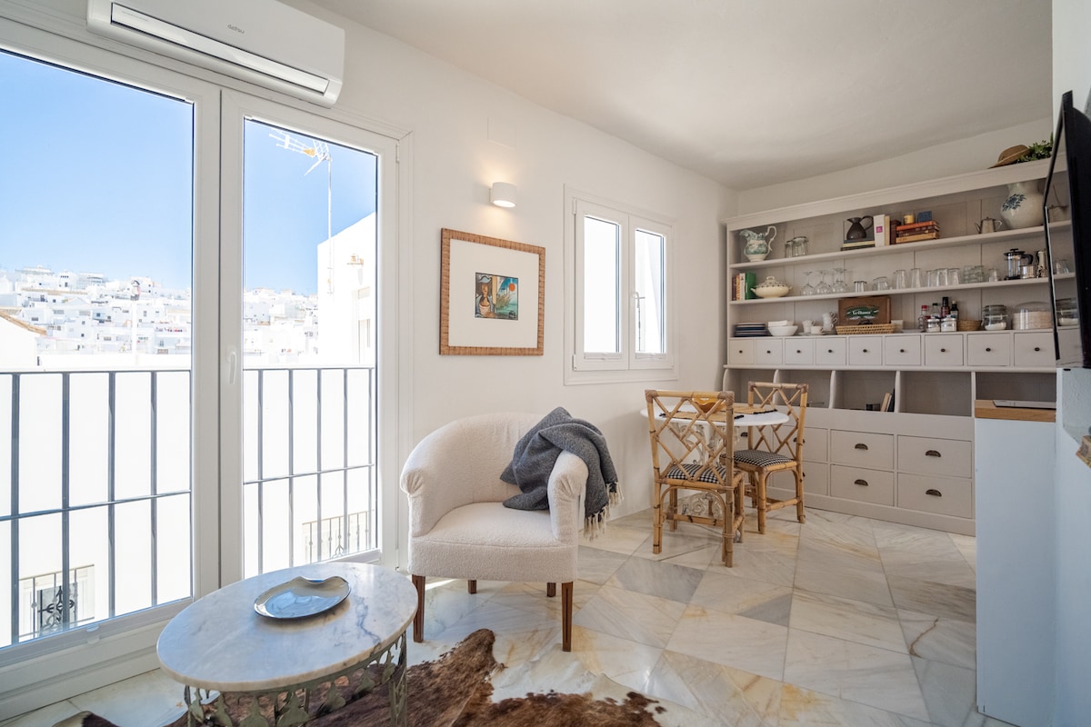A light-filled living space features large glass doors leading to a balcony, allowing natural light to enhance the room. A cozy armchair is paired with a round table and chairs, while shelves display decorative items. The polished marble floor adds elegance to the area.