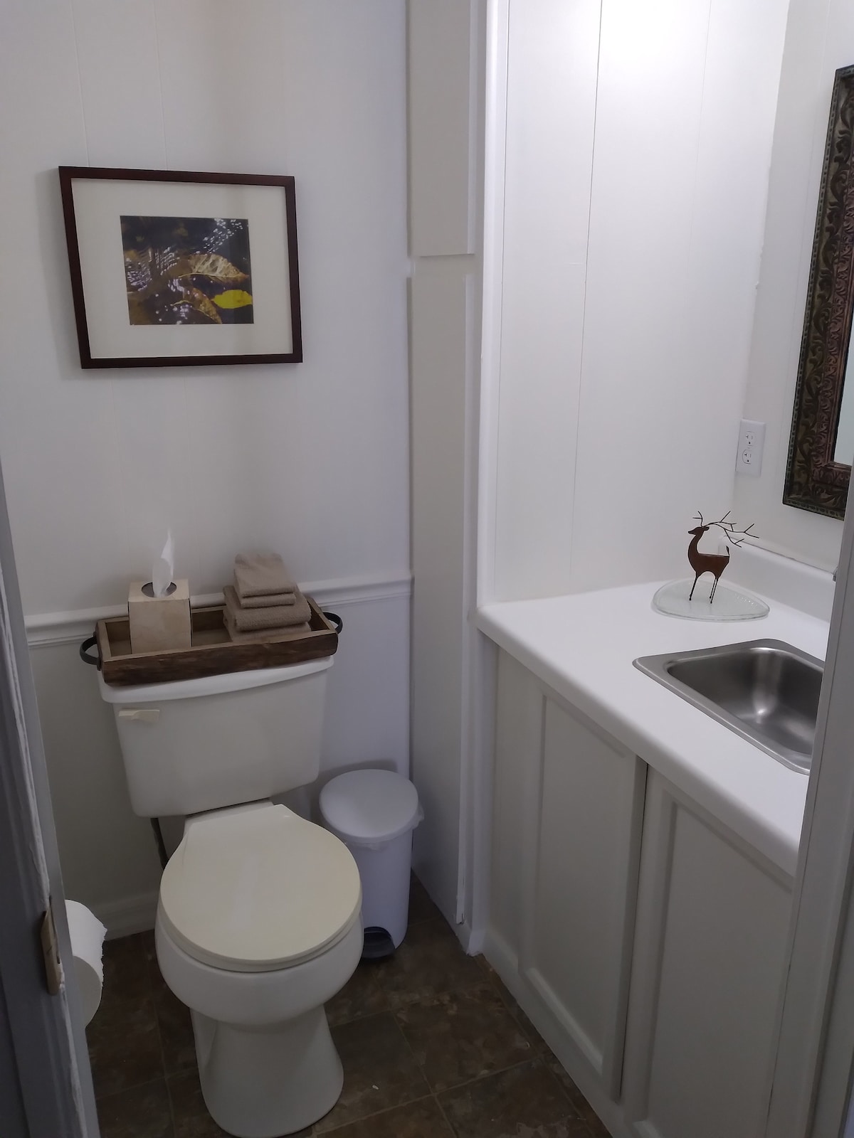 An elegant half bath features a compact design with a white toilet and a small sink area. Decorative towels are neatly arranged on a wooden tray. A framed artwork adorns the wall above the toilet, while a small decorative item sits on the counter.