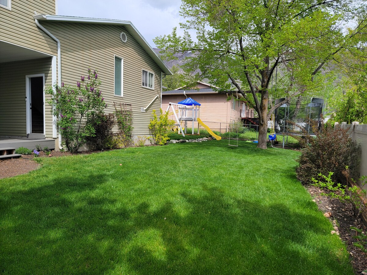 A green backyard is framed by a well-maintained lawn, featuring a slide and swingset in the background. Various plants and trees provide shade, while a trampoline is visible beyond the fence. The house structure displays neutral siding, and a paved path leads to the entrance.