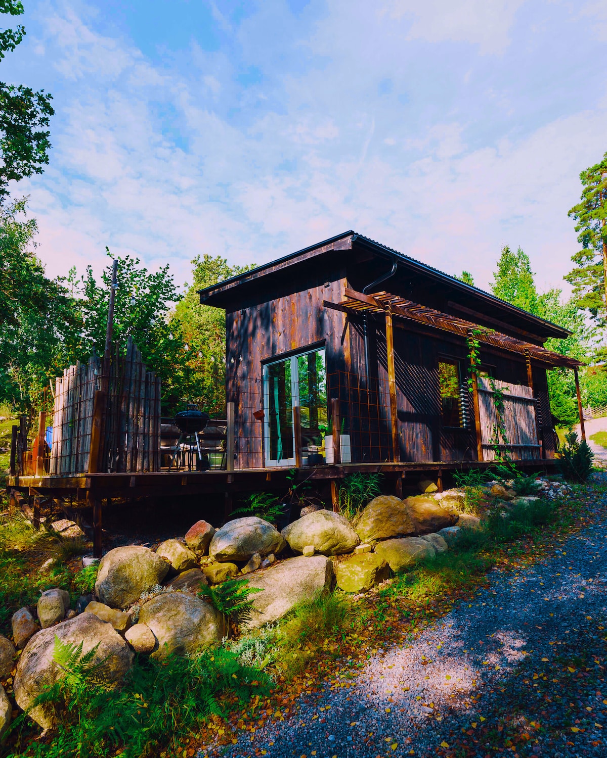 A cabin made of dark wood is set among greenery, showcasing a spacious outdoor deck with seating. Smooth stones line the path leading up to the structure. Large windows reflect natural light, inviting the surrounding landscape inside.