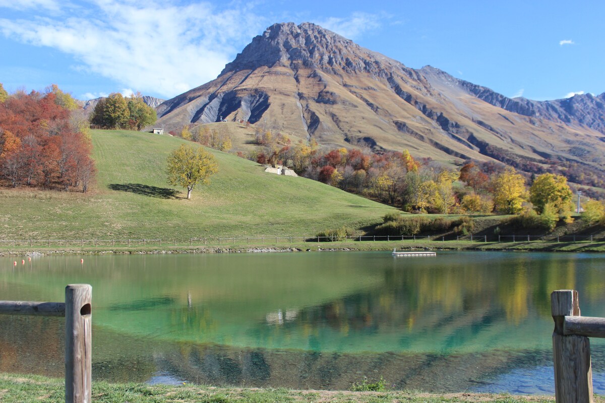 A serene landscape features a clear lake reflecting the surrounding hills. Autumn foliage paints the scene with warm colors, while a prominent mountain looms in the background. The tranquil environment is enhanced by gentle slopes and grassy areas, inviting peaceful outdoor exploration.
