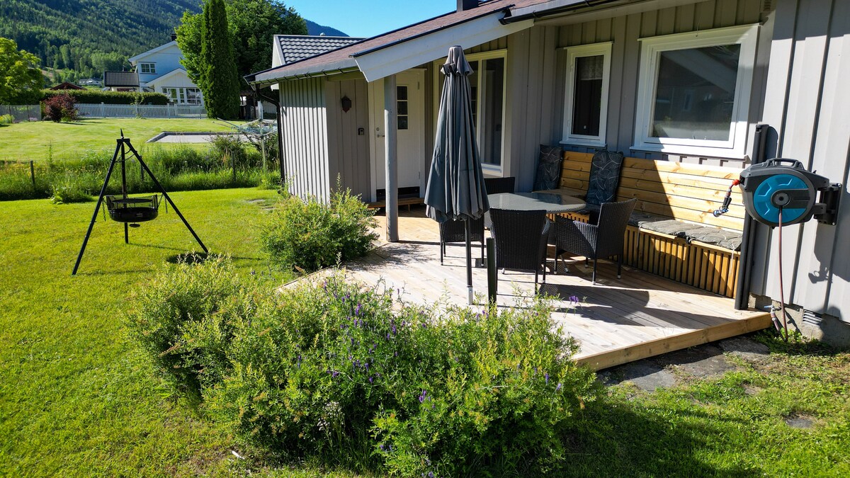 A wooden deck features a seating area with dark wicker chairs and a table, complemented by a large umbrella. A swing set is positioned nearby, surrounded by lush green grass and shrubs. Scenic mountains can be seen in the distance.