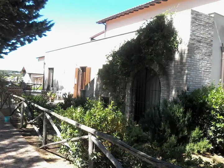 A spacious exterior view of the residence is presented, featuring a stone wall adorned with greenery. Wooden shutters frame the windows, while a wooden fence runs along the pathway. The landscape includes various shrubs and plants, enhancing the natural surroundings.