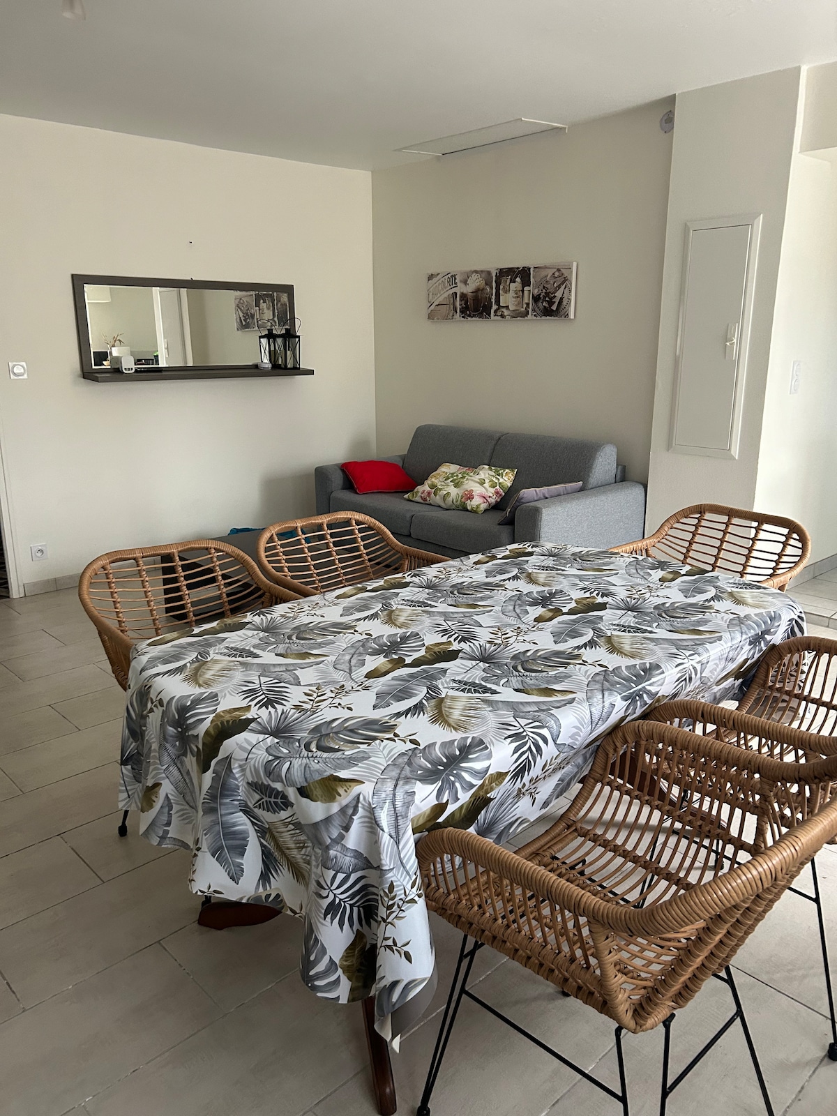 A dining table is set with a patterned tablecloth, surrounded by four wicker chairs. A gray sofa with colorful cushions is positioned in the background, accompanied by a wall-mounted mirror reflecting the natural light of the space.