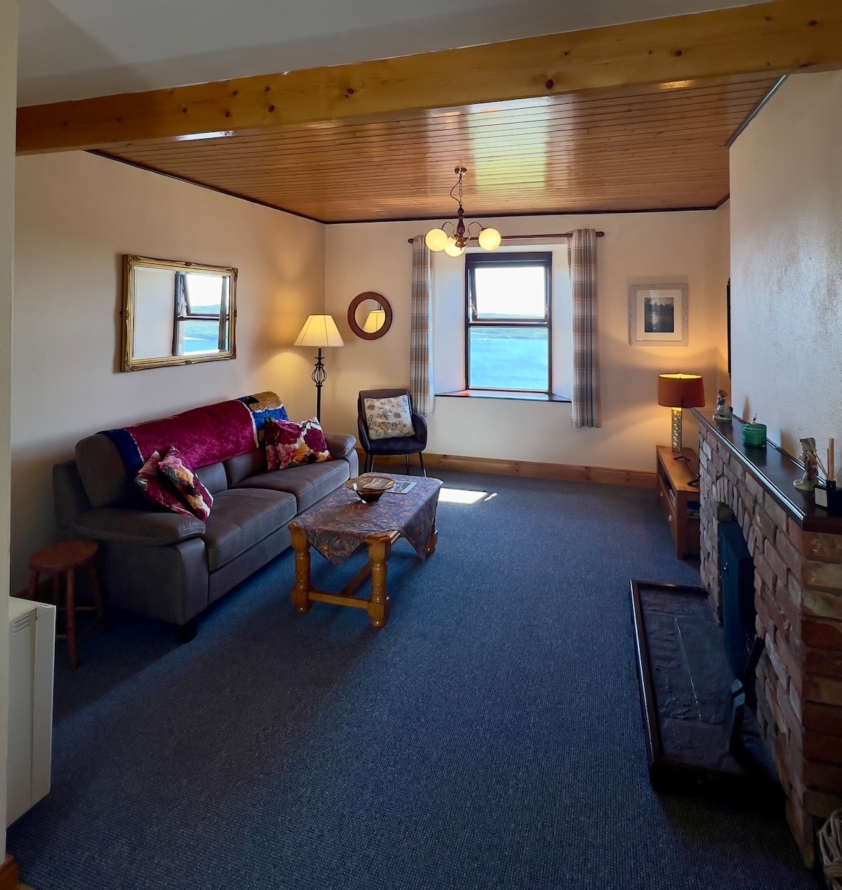 An open living area features a comfortable couch adorned with decorative pillows, a wooden coffee table, and a rustic brick fireplace. Natural light filters through a window adorned with plaid curtains, highlighting the warm wooden ceiling and wall decor.