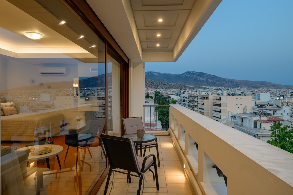 A private terrace with modern outdoor furniture overlooks a scenic view of the surrounding area. Large glass doors provide natural light to the interior space, where a cozy seating arrangement can also be seen. Soft lighting adds a warm ambiance to the setting.