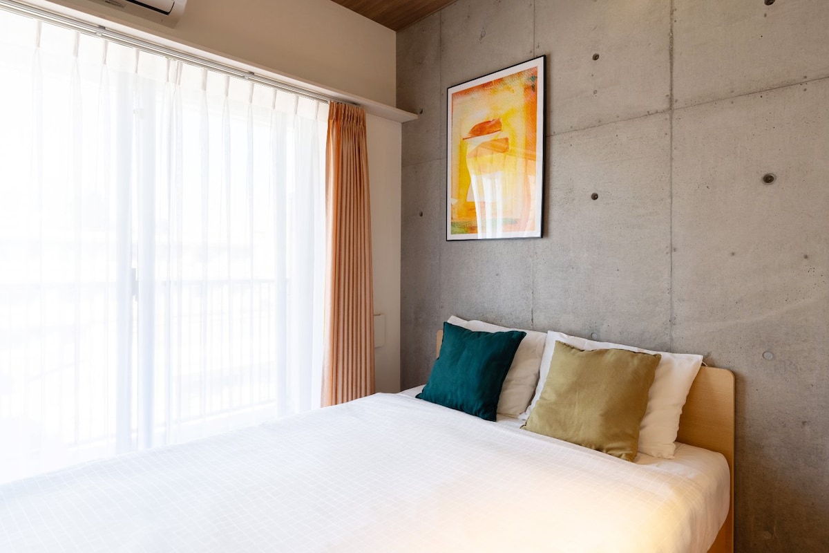 A cozy sleeping area is presented with a spacious bed adorned with decorative pillows in green and gold shades. Large windows are partially covered by sheer curtains, allowing natural light to illuminate the room. A colorful artwork hangs on the concrete wall, adding a touch of design.