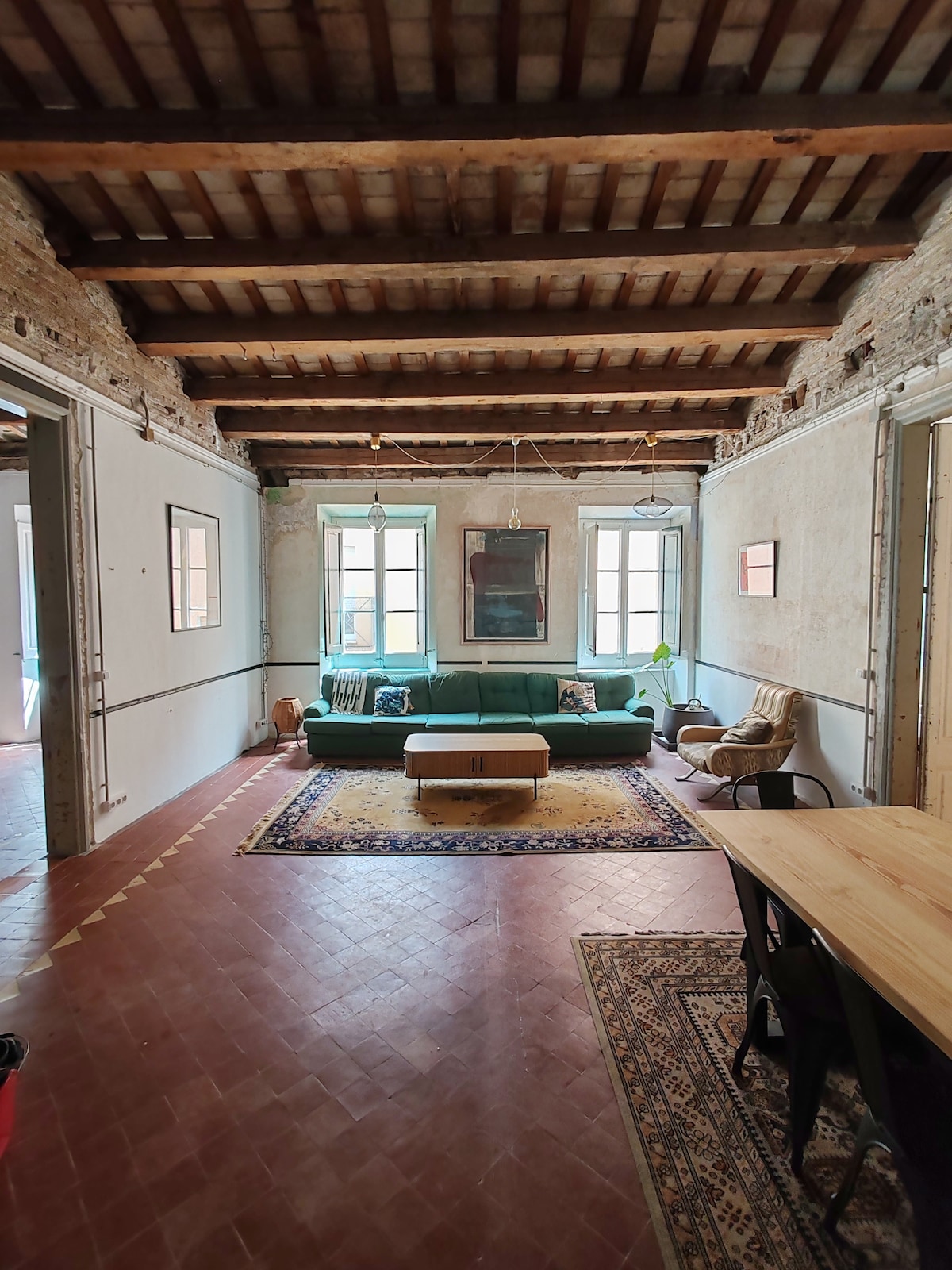 A spacious living area is highlighted by a green sofa, complemented by a colorful rug and central coffee table. Exposed wooden beams frame the ceiling, while natural light enters through multiple windows, creating an inviting ambiance. A wooden dining table is visible in the foreground.
