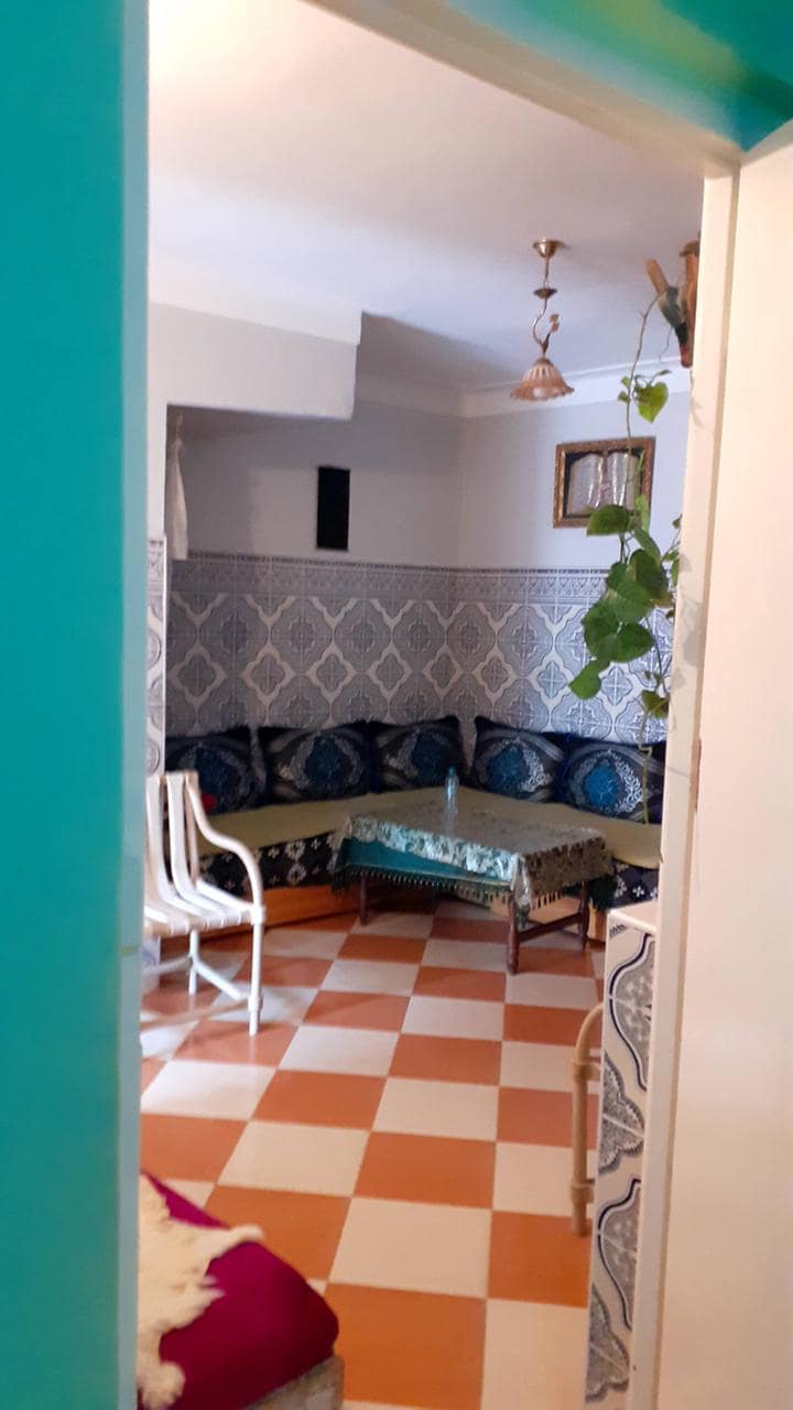A cozy seating area is visible through an open door, featuring patterned tiles in orange and white. A corner sofa with blue cushions surrounds a low table, and a potted plant adds a touch of greenery to the space.