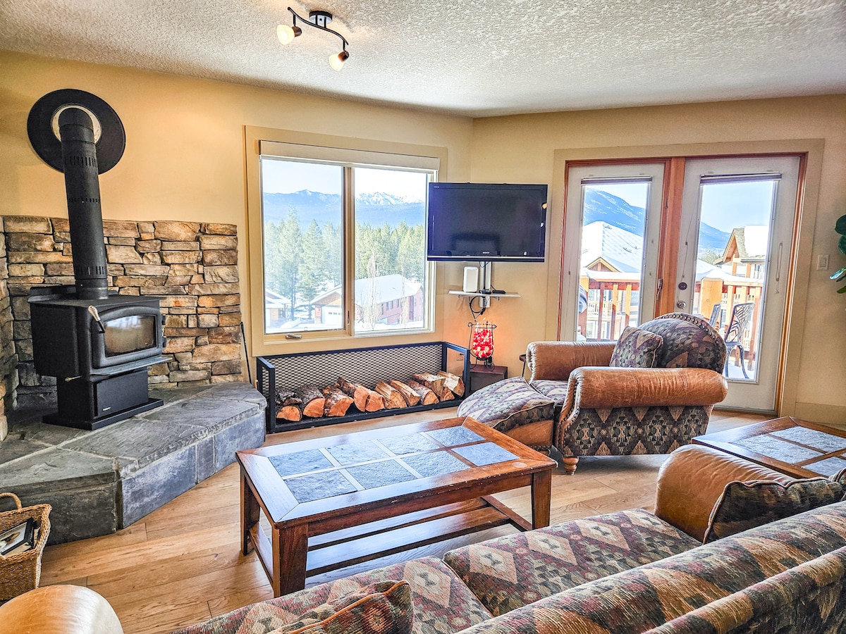The living area features a cozy wood-burning stove and a flat-screen TV. Large windows provide mountain views, while double doors lead to the outdoor balcony. A comfortable armchair and a wooden coffee table are included, enhancing the inviting atmosphere.