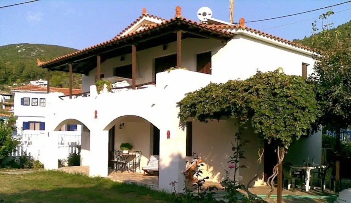 A traditional white building features a terracotta-tiled roof and wooden balconies. Lush greenery frames the entrance and outdoor seating area, with a small patio adorned with plants. The surrounding environment showcases a peaceful, garden-like space, inviting relaxation.