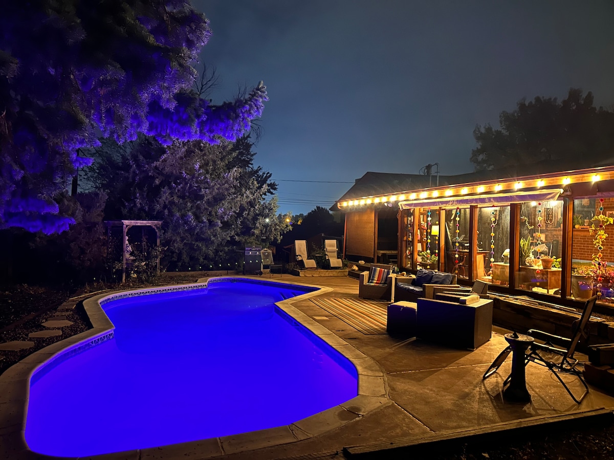 A well-lit outdoor area features a shimmering swimming pool, illuminated in blue hues. Lounge chairs are positioned nearby, with a cozy seating arrangement visible on a wooden deck, surrounded by lush greenery and trees. The inviting ambiance suggests a perfect space for relaxation.
