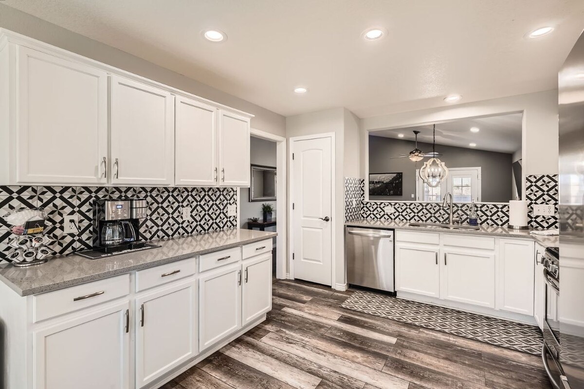 A modern kitchen is displayed, featuring white cabinetry and contrasting black and white tile backsplash. Stainless steel appliances are visible, and ample counter space is highlighted, with a cozy dining area seen through the doorway. Soft lighting enhances the welcoming atmosphere.