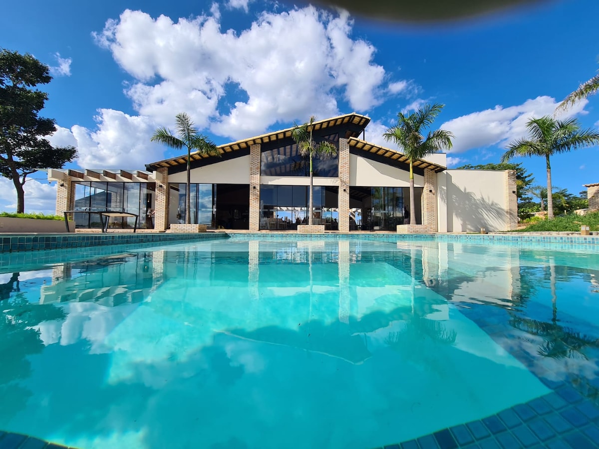 A modern structure with large glass windows reflects in the clear water of a spacious pool. Palm trees frame the view, and a serene sky filled with fluffy clouds adds to the atmosphere.