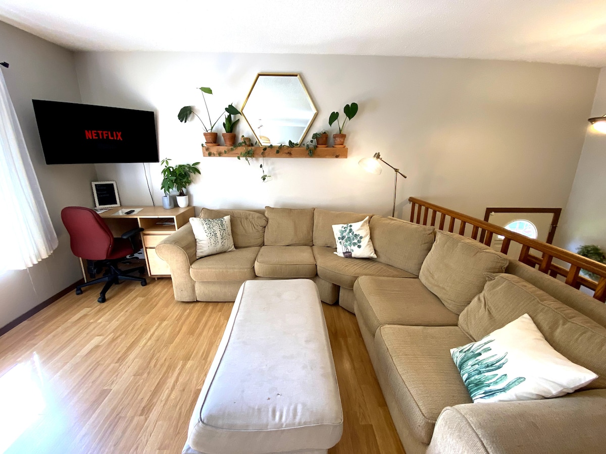 A spacious living area is furnished with a large sectional sofa and a rectangular coffee table. A flat-screen television is mounted on the wall, accompanied by a small desk and an office chair. Green plants and decor elements are displayed on a shelf above the couch.
