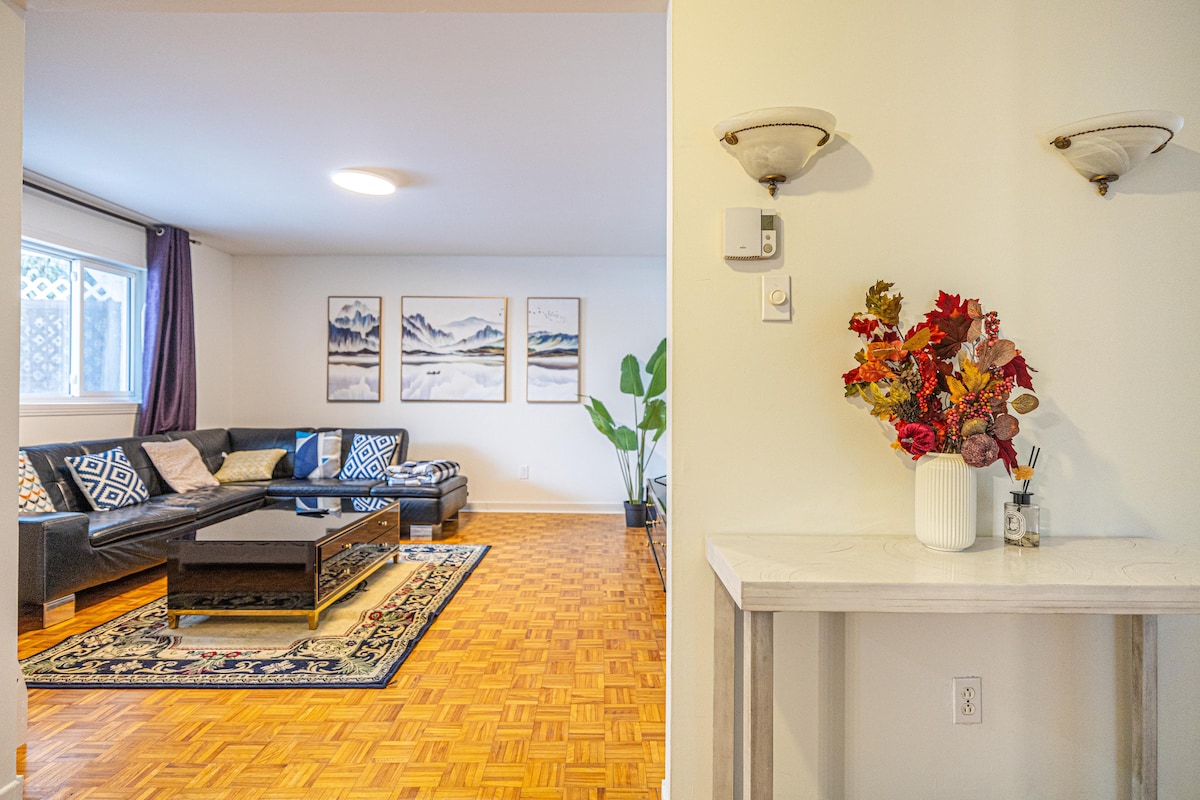 A spacious living area is revealed with a large sectional sofa facing a coffee table. Artwork depicting mountainous landscapes decorates the wall, while a potted plant adds a touch of greenery. A small side table holds a vase filled with decorative flowers, enhancing the welcoming ambiance.