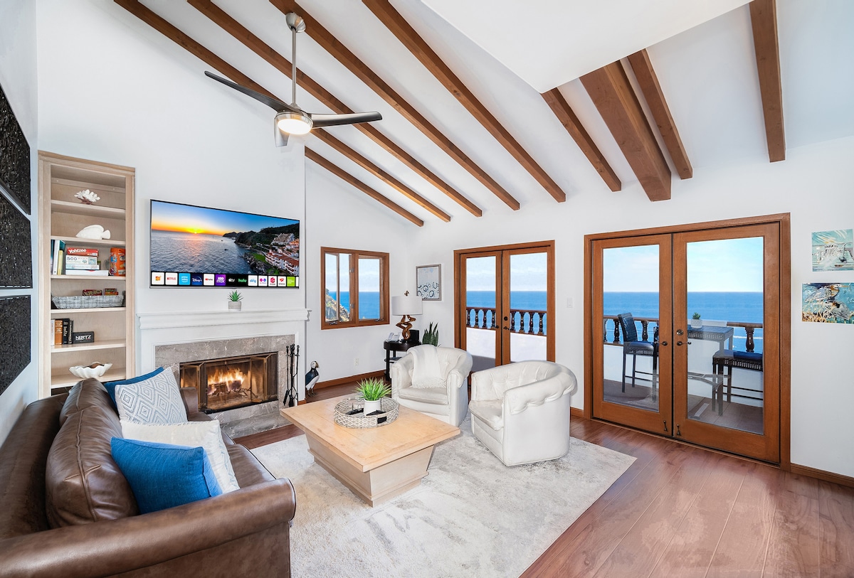 The living room features high, vaulted ceilings with exposed beams. A comfortable leather sofa and two armchairs are centered around a wooden coffee table. A modern fireplace is located on one side, while large French doors open to ocean views. A wall-mounted TV displays various media options.