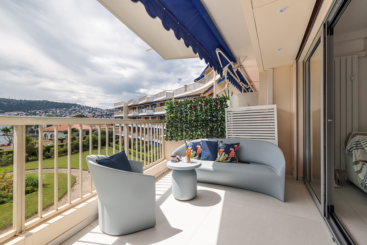 A spacious terrace features comfortable seating arrangements, including a curved sofa and a small table. Potted plants and greenery create a sense of privacy, while an awning provides shade. Views of the surrounding landscape and distant hills are visible in the background.