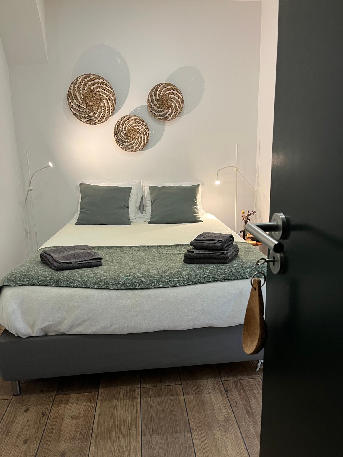 A softly lit bedroom showcases a spacious bed topped with gray pillows and a green throw blanket. Neatly folded towels are arranged at the foot of the bed. Woven circular wall decor adds texture to the minimalistic design, with light fixtures providing warm illumination.