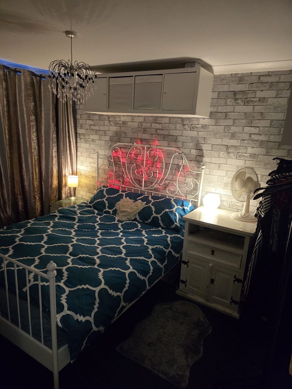 A cozy sleeping area features a white metal bed with a blue patterned duvet and decorative pillows. A stylish lamp and a fan are positioned on a nightstand, while soft light streams from a nearby window covered by elegant curtains. Decorative wall paneling adds character to the space.