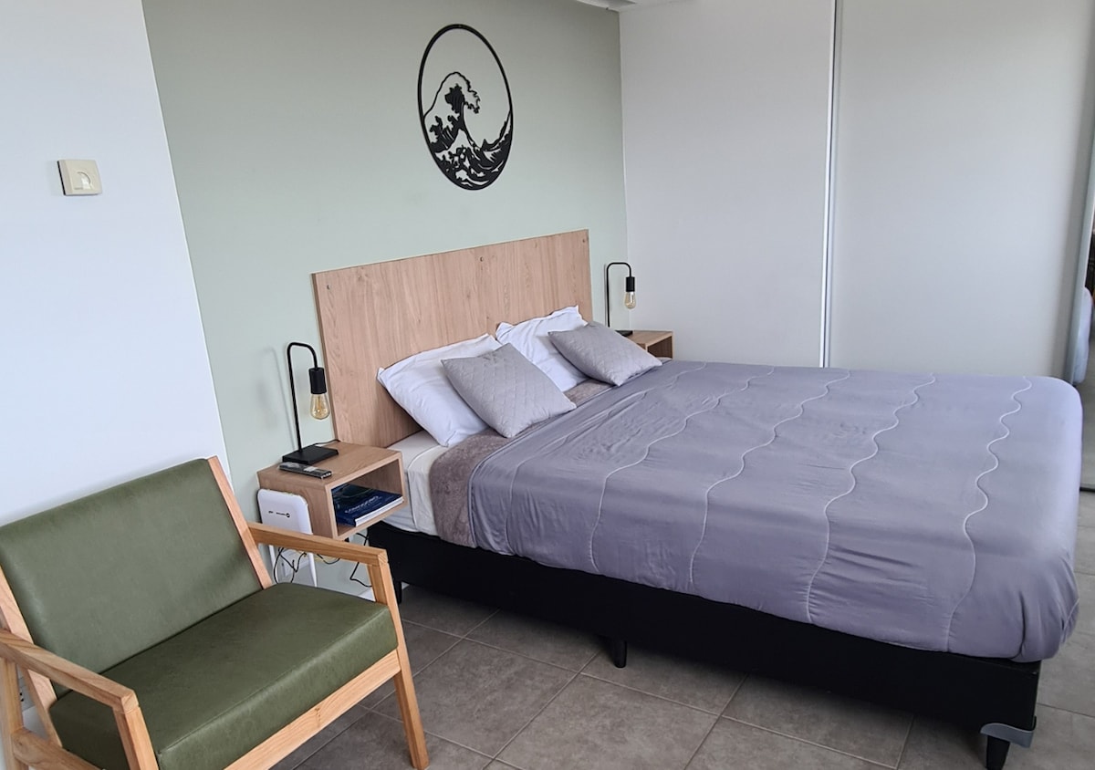 A cozy sleeping area is equipped with a spacious bed adorned with neutral linens. A wooden headboard complements the light green wall, while a green armchair adds a touch of color. Soft lighting fixtures on both sides enhance the room's functionality.