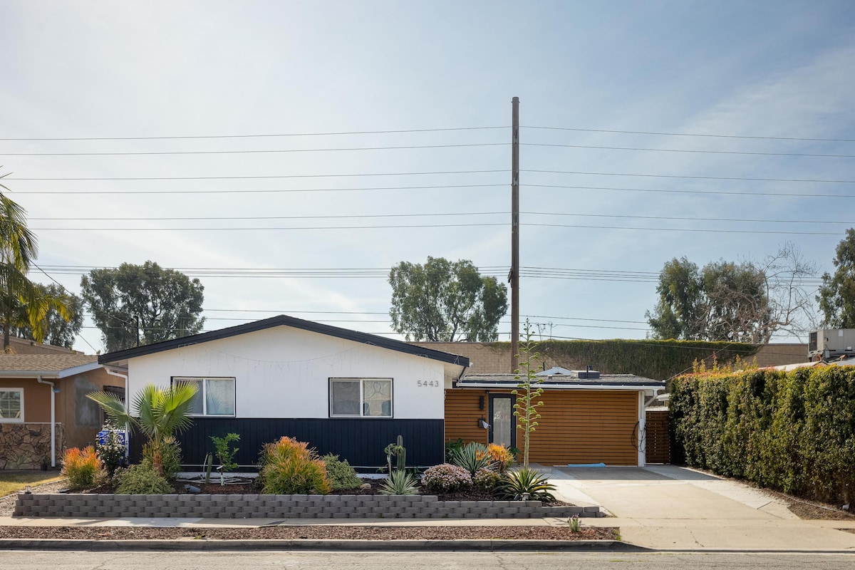 A single-story home is presented with a monochromatic exterior, featuring contrasting elements of white and dark blue. Lush greenery and low-maintenance landscaping frame the property, while a paved driveway provides convenient access. The surrounding area shows a blend of residential architecture and foliage.