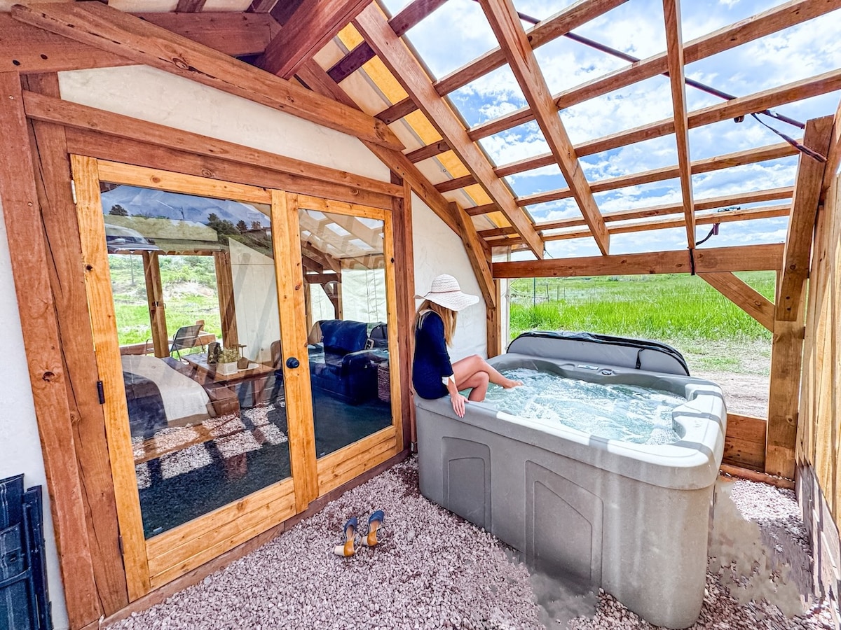 A shaded area with a hot tub is visible beside large glass doors leading into the tent interior. Light filters through the wooden beams above, illuminating a cozy space with comfortable seating. A person is seated at the edge of the hot tub, enjoying the serene surroundings.