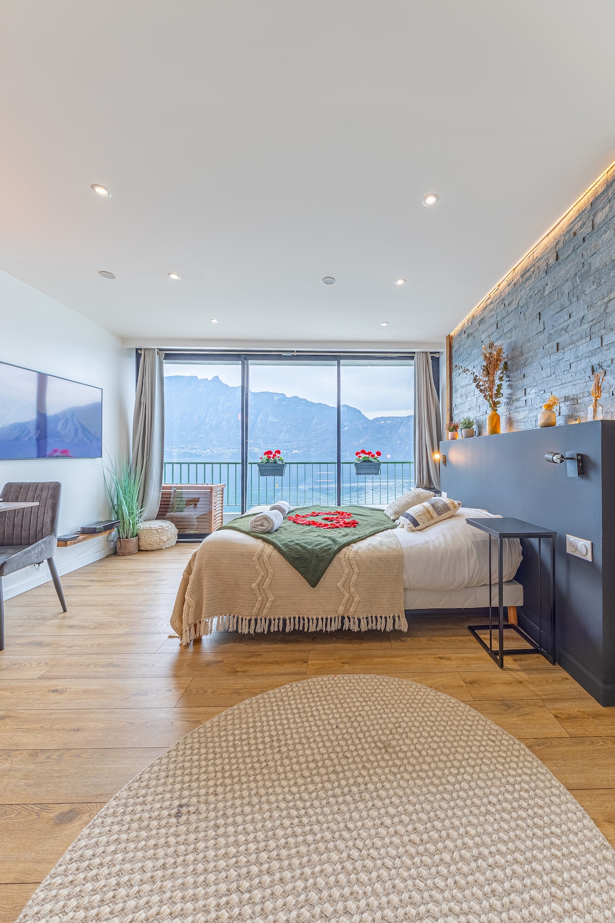 A bright bedroom features a spacious bed with a cozy blanket. Expansive windows showcase a stunning mountain view, while modern decor includes a small side table and plants. Natural light enhances the wood flooring, creating a warm atmosphere.