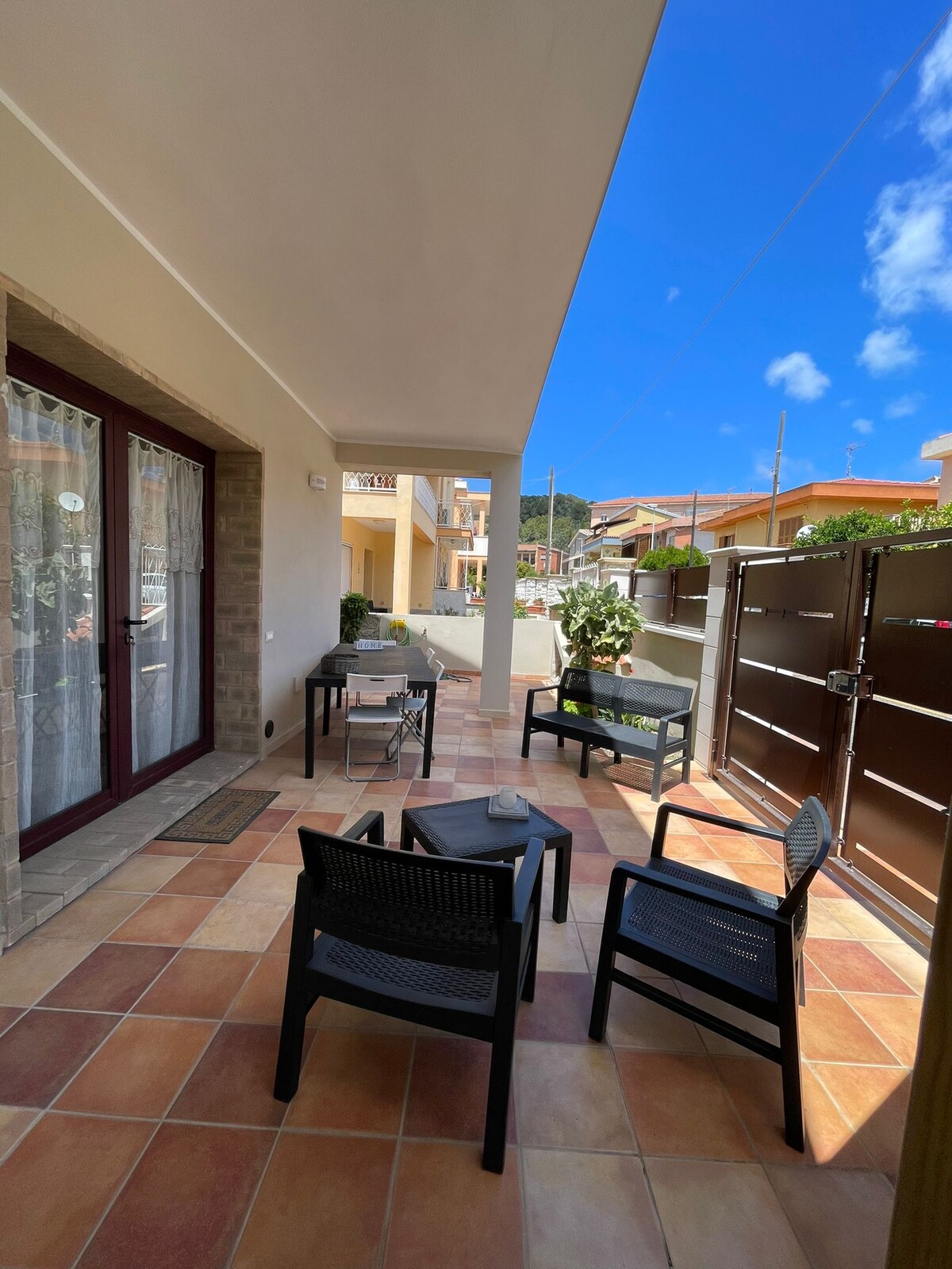 A covered patio area is equipped with modern, dark-colored furniture, featuring four chairs surrounding a small table. Doors lead into the interior, and natural light illuminates the space through high walls. The sky above is clear and bright, suggesting a pleasant outdoor environment.