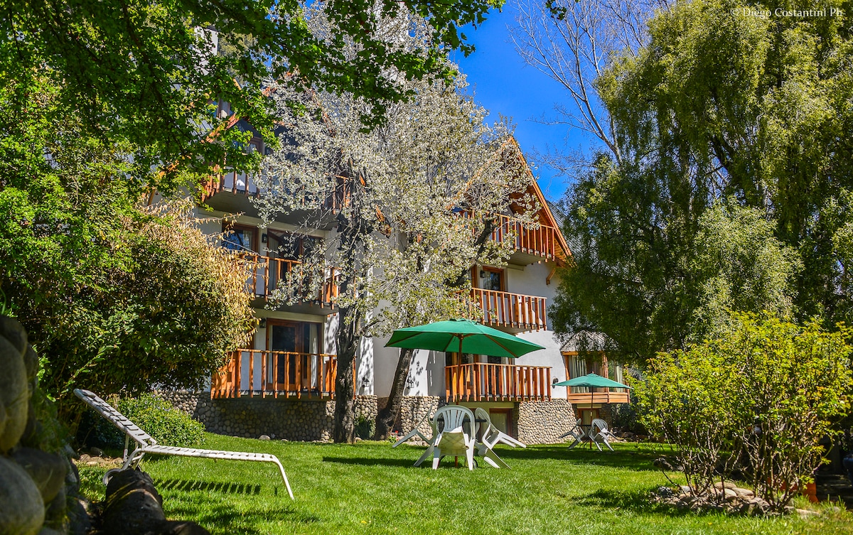 A scenic view of the apartment complex, framed by lush greenery and flowering trees. The exterior features multiple balconies with wooden railings, and a spacious lawn is adorned with lounge chairs and green umbrellas, creating a relaxing outdoor space.
