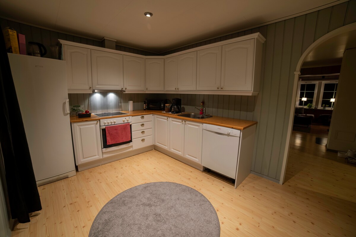 A modern kitchen features white cabinetry and wooden countertops. Essential appliances include a refrigerator, stove, and dishwasher. Bright lighting illuminates the space, enhancing the clean design. A circular rug is placed on the light wooden floor, creating a cozy atmosphere.