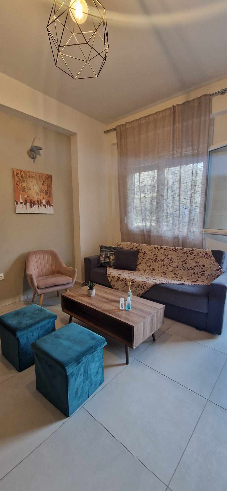 A comfortable seating area features a dark sofa adorned with decorative cushions. A wooden coffee table is centered between two soft ottomans in vibrant teal. An accent chair with light upholstery complements the space. Natural light enters through large windows framed by sheer curtains.