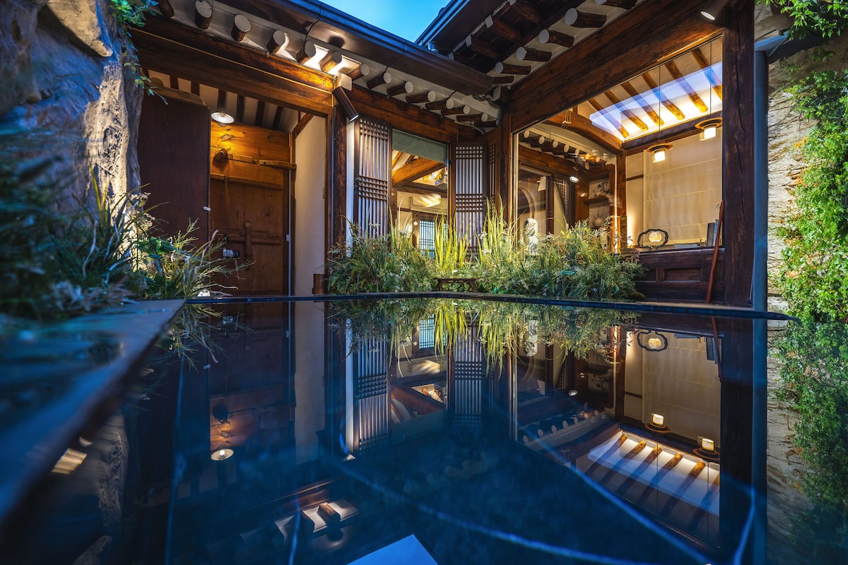 A tranquil outdoor space is captured, featuring a reflective pool surrounded by lush greenery. Traditional wooden structures are visible, showcasing architectural details. Soft lighting enhances the serene ambiance, creating a peaceful environment suitable for relaxation.