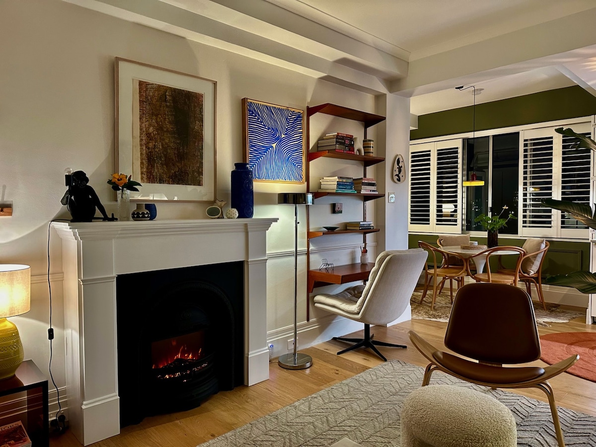 A cozy living space features a decorative fireplace with an electric insert. A white armchair and a brown chair are positioned nearby. Shelving displays various books and art, while a dining area with a round table and chairs is visible through large windows adorned with plantation shutters.