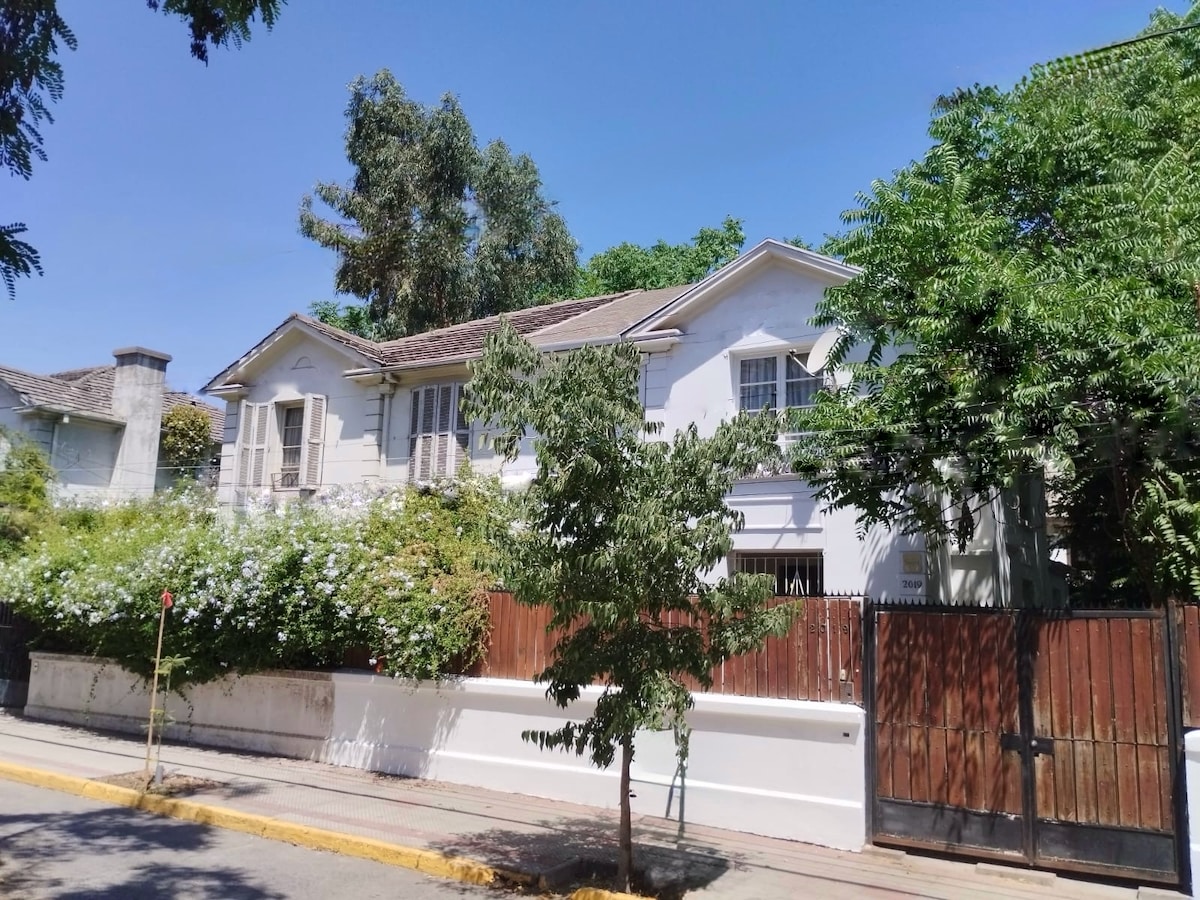 The exterior of a renovated two-story house is showcased, featuring white walls and large windows framed by wooden shutters. Lush greenery surrounds the property, enhancing its curb appeal. A wooden fence offers privacy while a tree adds a touch of nature to the front yard.