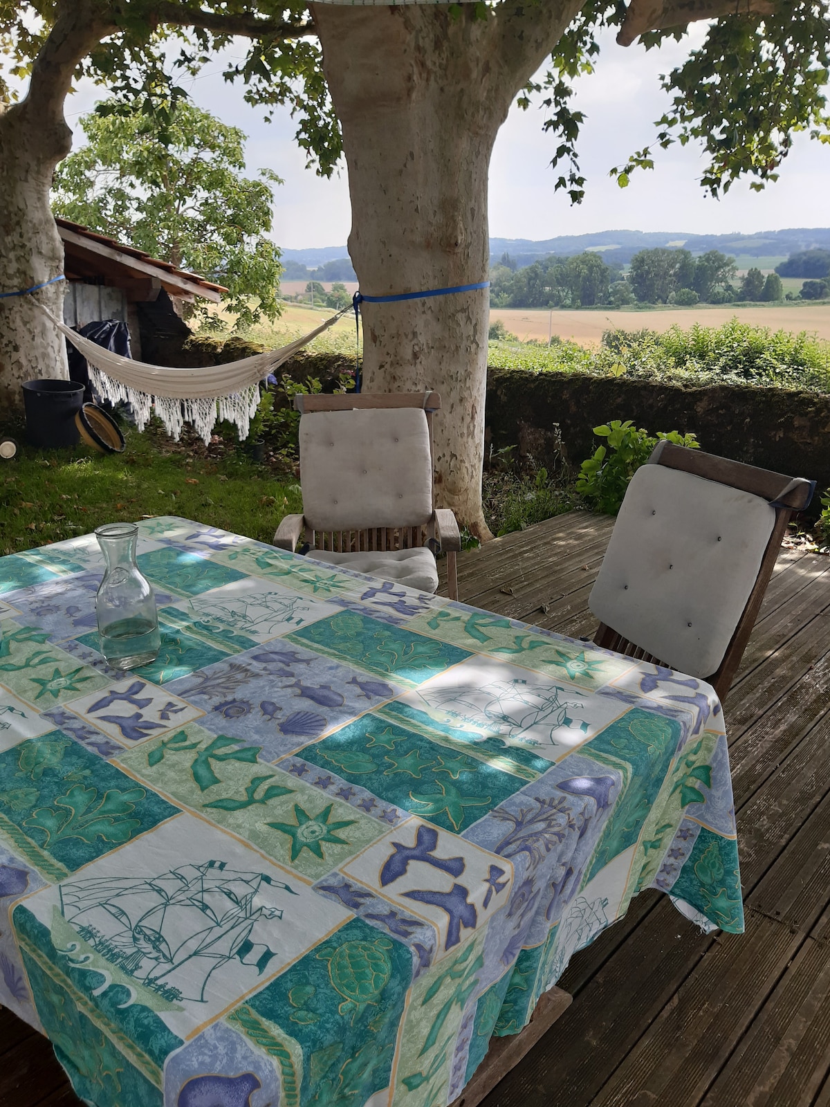 A shaded outdoor area features a table covered with a patterned cloth, accompanied by two comfortable chairs. A hammock is seen nearby, with lush greenery and rolling hills visible in the background, providing a serene countryside view.