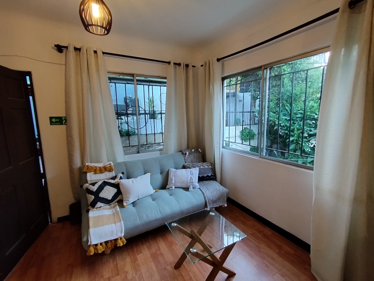 A cozy living area is presented, featuring a light blue sofa adorned with decorative pillows. A glass coffee table occupies the center space, while large windows frame views of greenery outside. Sheer curtains allow natural light to fill the room, enhancing its inviting ambiance.