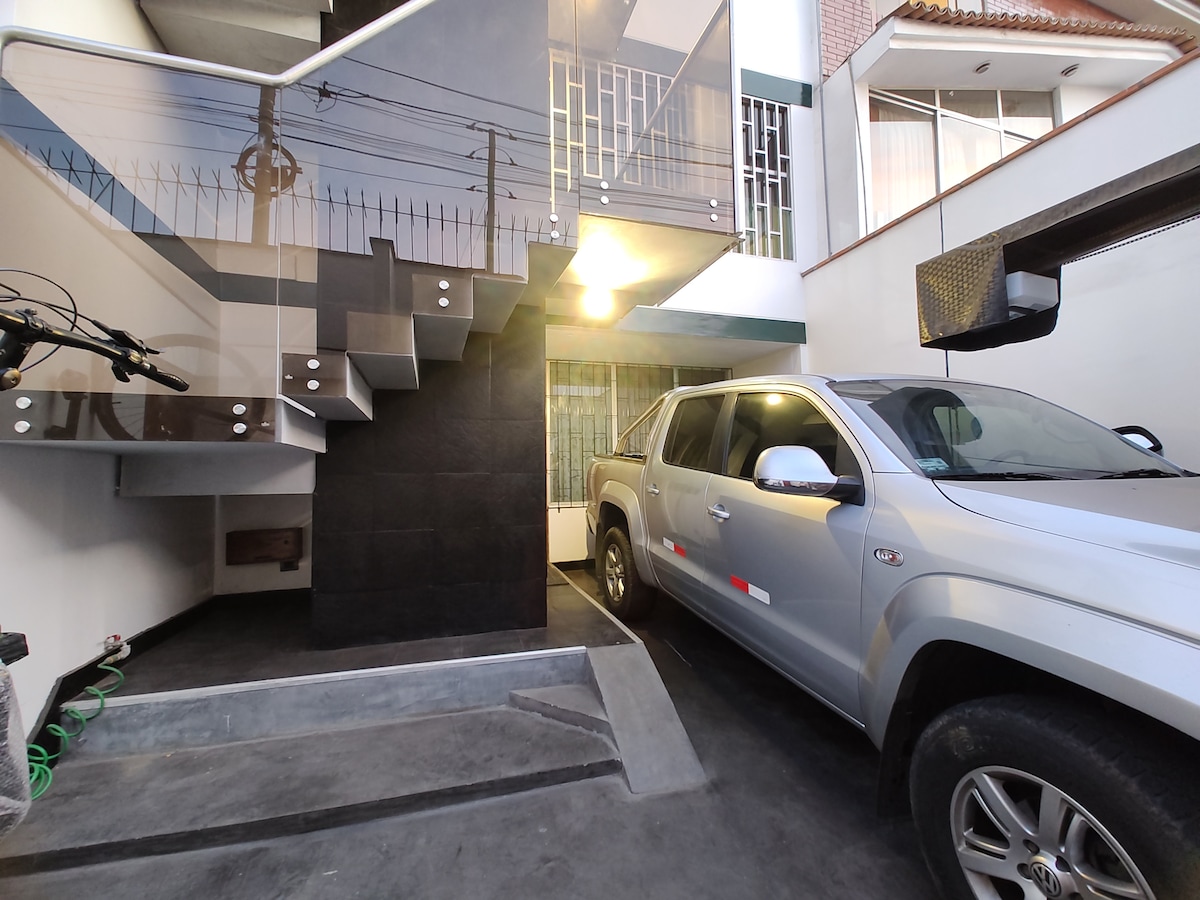 A secure parking area is presented with a grey vehicle parked underneath a staircase. The space features tiled flooring and walls that create a modern aesthetic. Natural light is seen illuminating the area from a nearby window, providing a sense of openness.