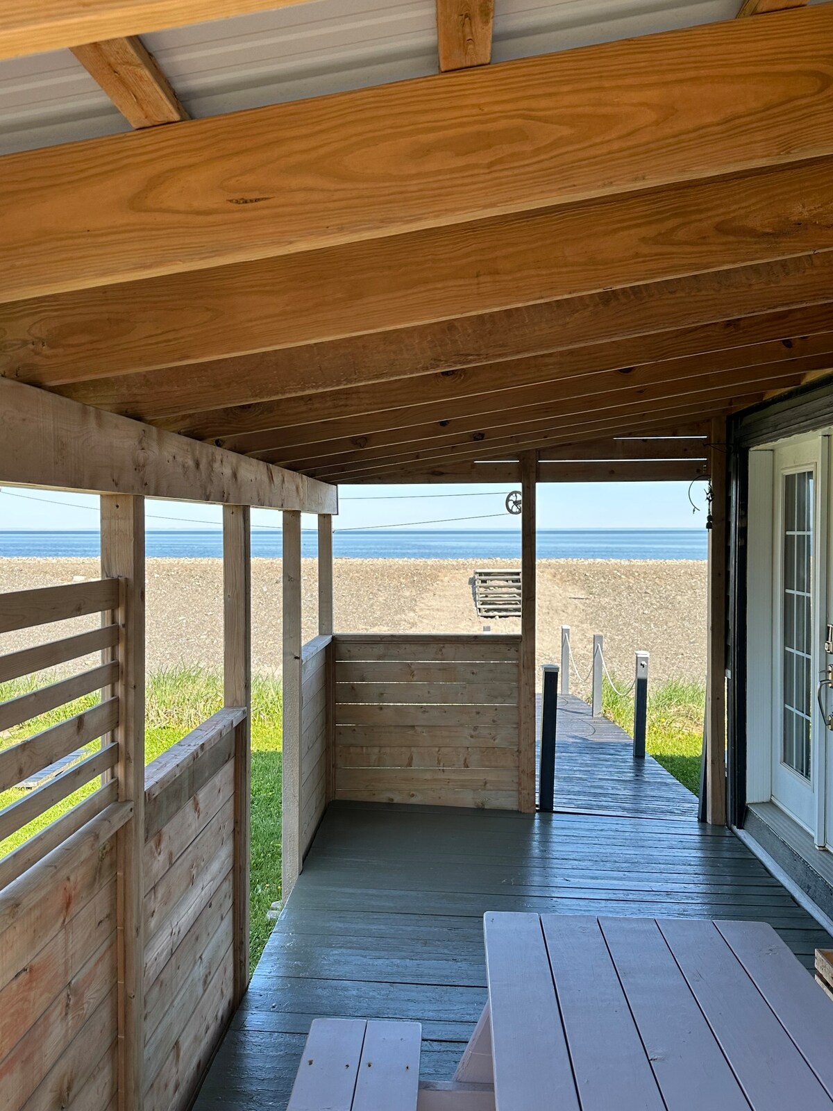 A covered terrace features wooden walls and a spacious table, offering a shaded area for dining. The view extends towards a gravel beach and the open sea beyond, with a pathway leading from the terrace towards the water.