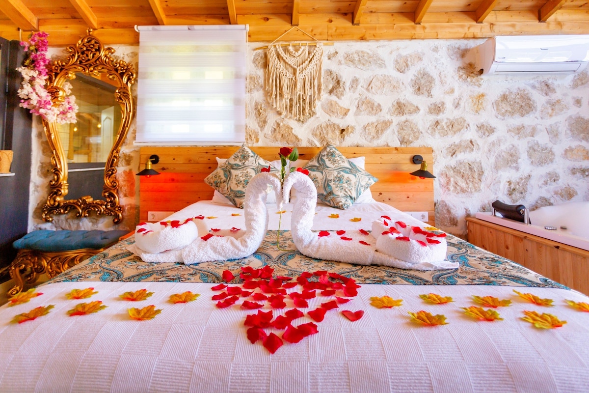 A cozy sleeping area features a bed adorned with towel sculptures resembling swans, surrounded by scattered rose petals. Decorative pillows are positioned against a wooden headboard. A wooden mirror with ornate detailing is visible, complemented by soft lighting from a nearby window.