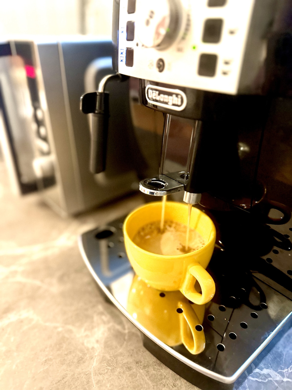 A coffee machine is positioned on a sleek countertop, pouring freshly brewed coffee into a bright yellow cup. The machine's control panel features dials and buttons, while reflections and coffee drips can be seen on the polished surface.