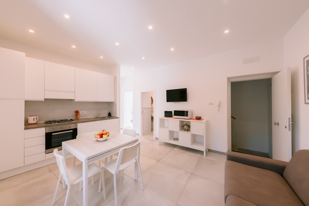 A modern kitchen space is highlighted, featuring white cabinetry and stainless steel appliances. A white dining table with chairs is positioned centrally, complemented by a cozy sofa in a neutral tone. Soft lighting illuminates the area, creating a bright and open atmosphere.