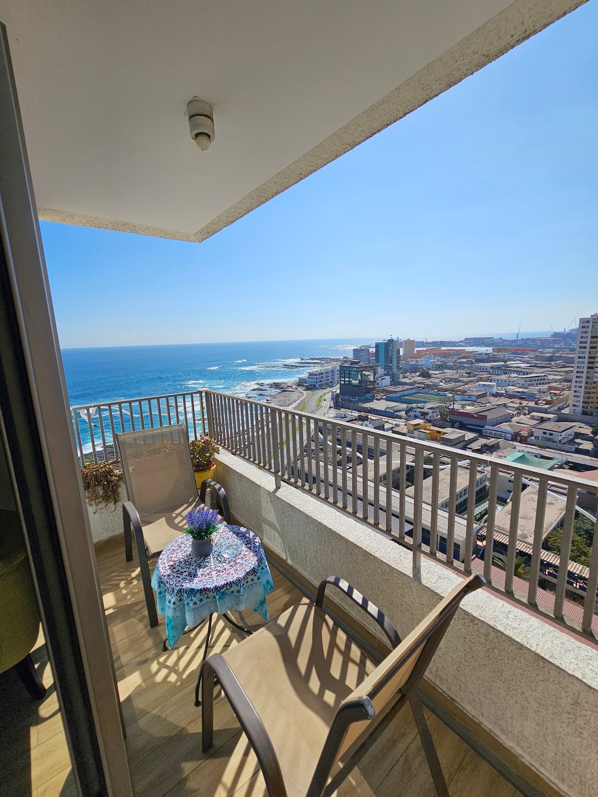 A small balcony is shown, featuring a round table draped with a blue tablecloth and a bouquet of flowers. Two chairs are positioned around the table, with a view overlooking the ocean and coastline in the background. Clear skies enhance the scenic landscape.