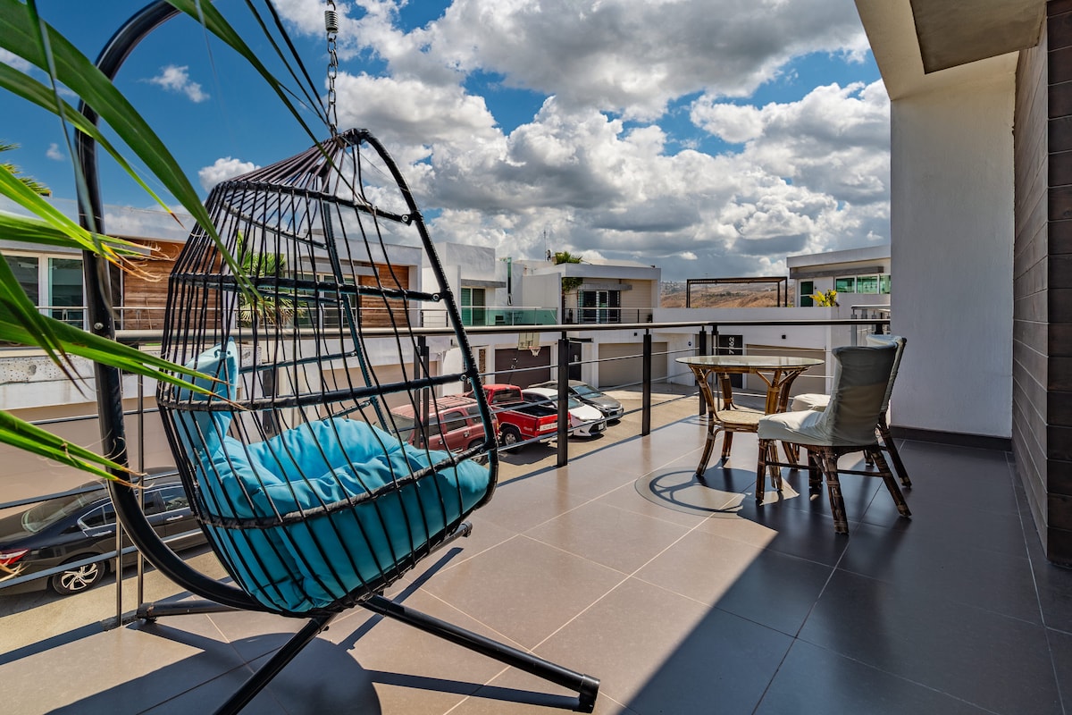 A modern balcony space features a suspended chair with blue cushions, offering a comfortable seating option. A small round table is accompanied by two chairs, all set against a backdrop of a bright sky with scattered clouds. The balcony overlooks a driveway area with parked cars.