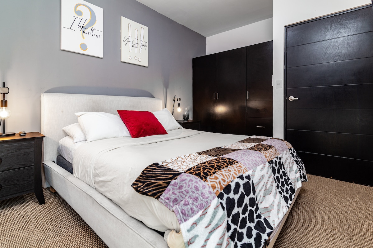 A spacious bedroom is presented with a light-colored bed featuring decorative throws in various textures and patterns. Two stylish wall art pieces are mounted above the bed, complementing the dark wooden cabinetry in the background. Warm bedside lighting enhances the cozy environment.