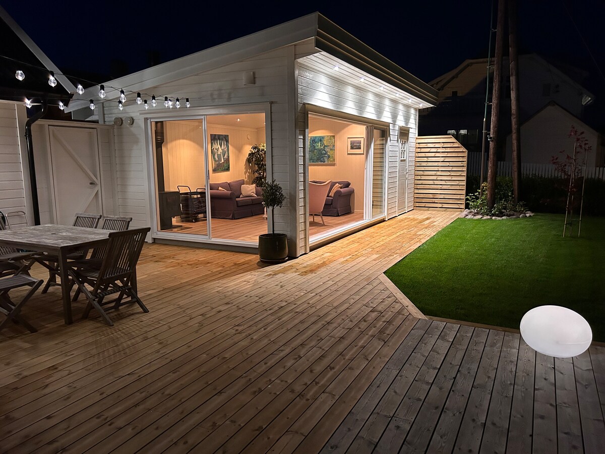 An inviting outdoor space is presented with wooden decking and a well-maintained lawn. The cozy living area is visible through expansive glass doors, allowing for a seamless transition between indoor and outdoor environments. Soft lighting enhances the tranquil atmosphere.