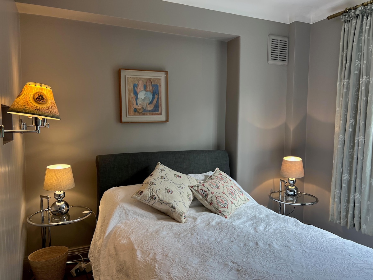 A cozy bedroom is presented, featuring a spacious bed with a white quilt and decorative pillows. Wall lamps provide soft lighting beside the bed. A framed artwork is hung above the headboard, while patterned curtains adorn the window, adding a touch of elegance to the neutral-toned walls.
