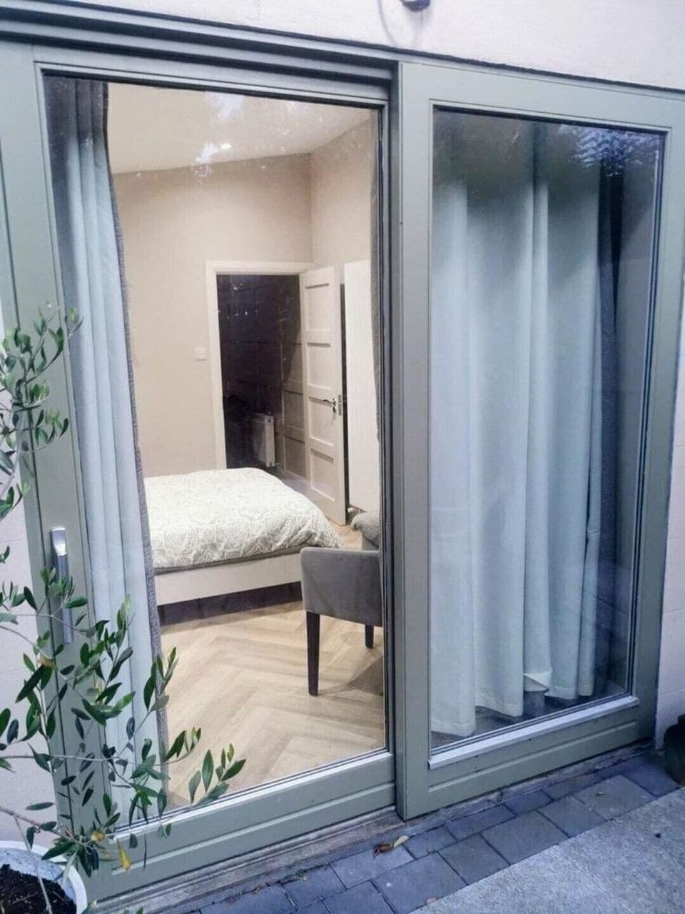 A private bedroom is visible through sliding glass doors, featuring a spacious bed with a light-colored coverlet. A soft chair is positioned nearby, and light-colored curtains frame the windows, allowing natural light to fill the room. A glimpse of a bathroom area is seen in the background.
