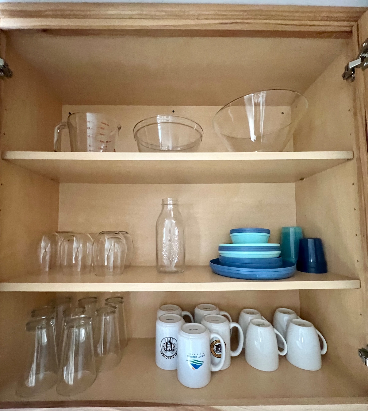 A wooden cabinet is filled with various glassware and dishware. The top shelf contains measuring cups and bowls, while the middle shelf features a mixture of glass and colorful plates. The bottom shelf displays an assortment of mugs and bottles, arranged neatly.