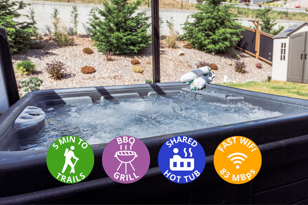 A covered hot tub is seen bubbling in a private backyard setting, surrounded by greenery. Soft towels are neatly arranged on the edge. The space provides opportunity for relaxation after exploring nearby trails.